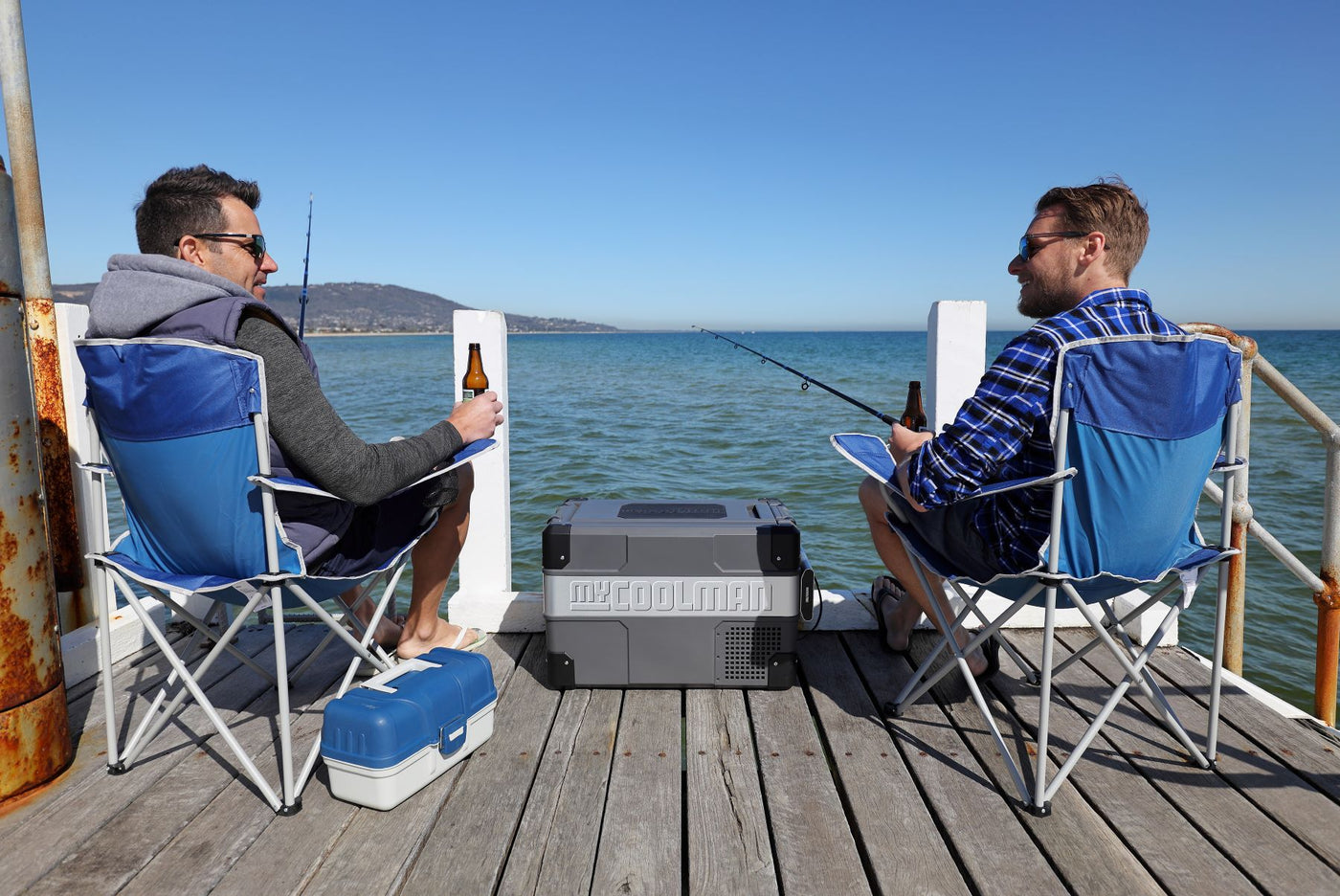 Two men sitting on chairs on a wharf holding fishing rods with myCOOLMAN fridge in the middle