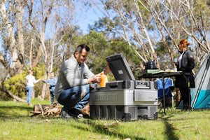 Man lifting orange juice out of a myCOOLMAN with people and camping equipment in background